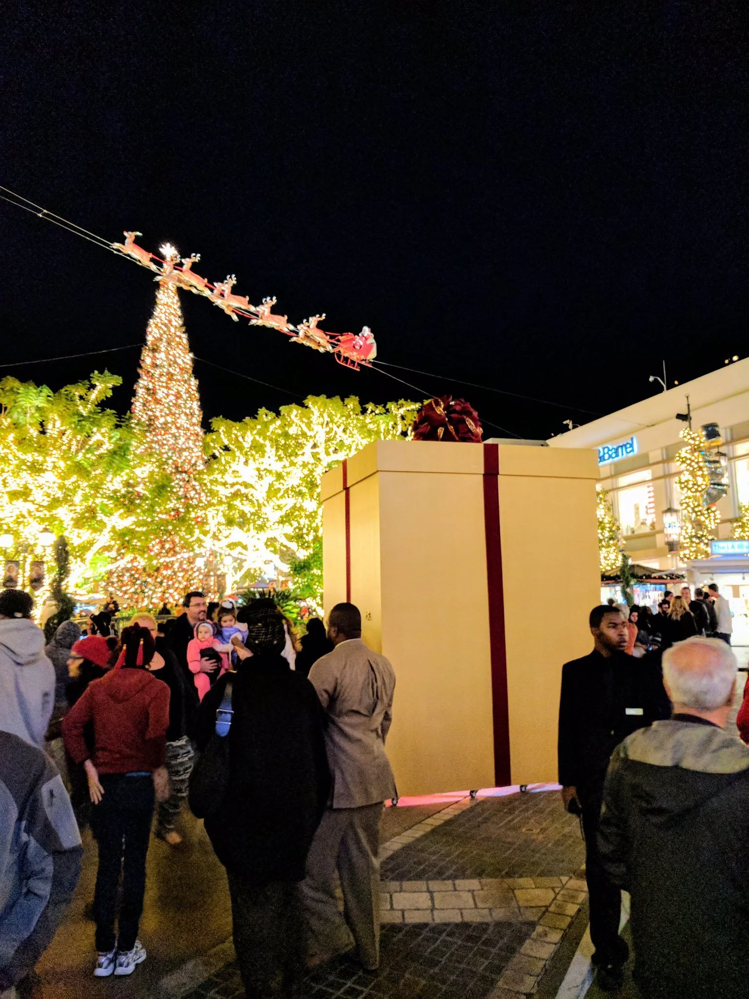 Crowd at night beside a giant gift-wrapped box installation, with a lit Christmas tree and Santa's sleigh overhead